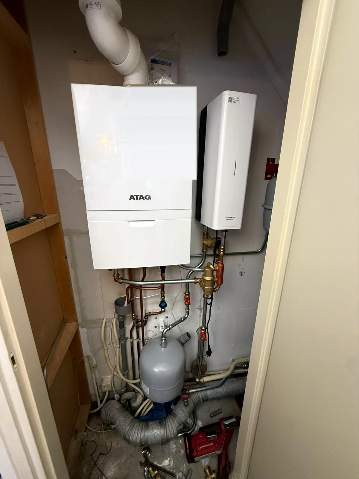 Atag cv-ketel met daarnaast een smalle witte boilerunit, verbonden met koperen en stalen leidingen boven een expansievat