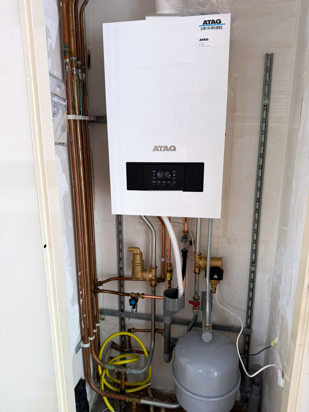 Atag cv-ketel in een smalle kast met koperen leidingen, een gele gasslang en een grijs expansievat eronder