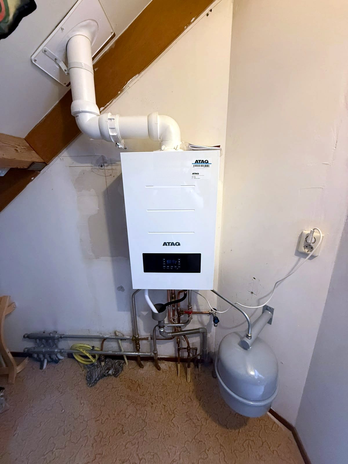 Witte Atag cv-ketel onder een schuine trap, met een wit rookgasafvoerkanaal langs de wand en een expansievat op een beugel ernaast