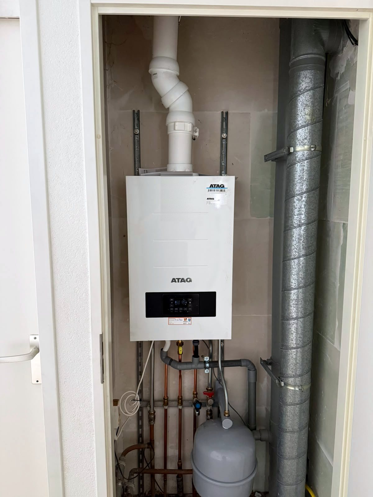 Atag cv-ketel in een technische kast naast een grijs geïsoleerd ventilatiekanaal, met leidingen en een expansievat onder de ketel