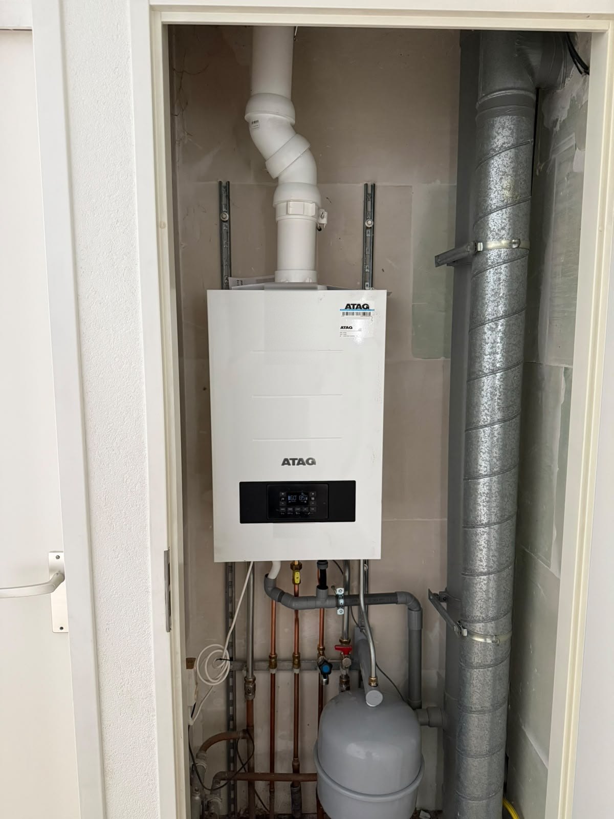 Atag cv-ketel in een technische kast naast een grijs geïsoleerd ventilatiekanaal, met leidingen en een expansievat onder de ketel
