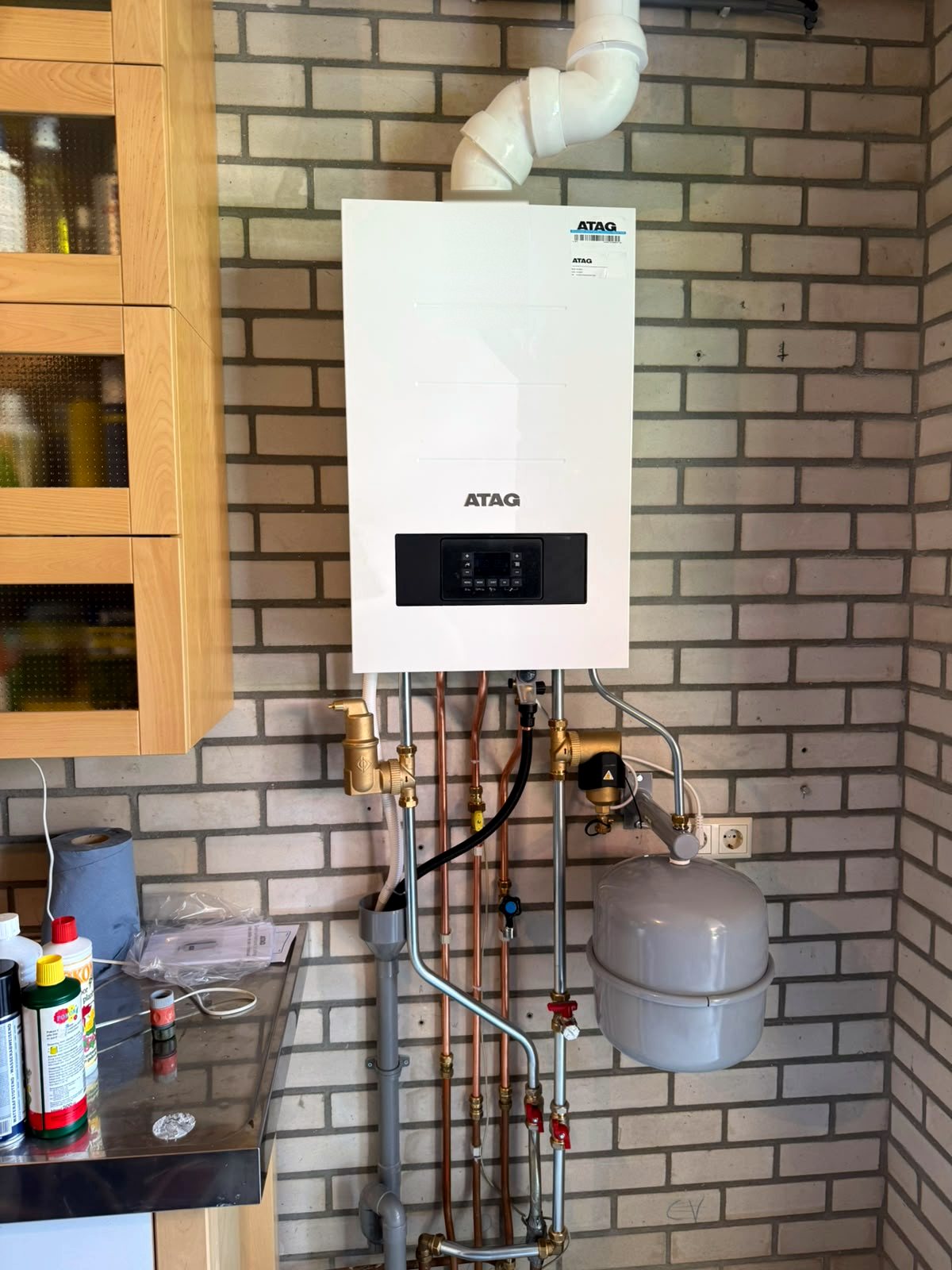 Atag cv-ketel tegen een lichte bakstenen wand in een bijkeuken, met koperen leidingen en een grijs expansievat aan de muur