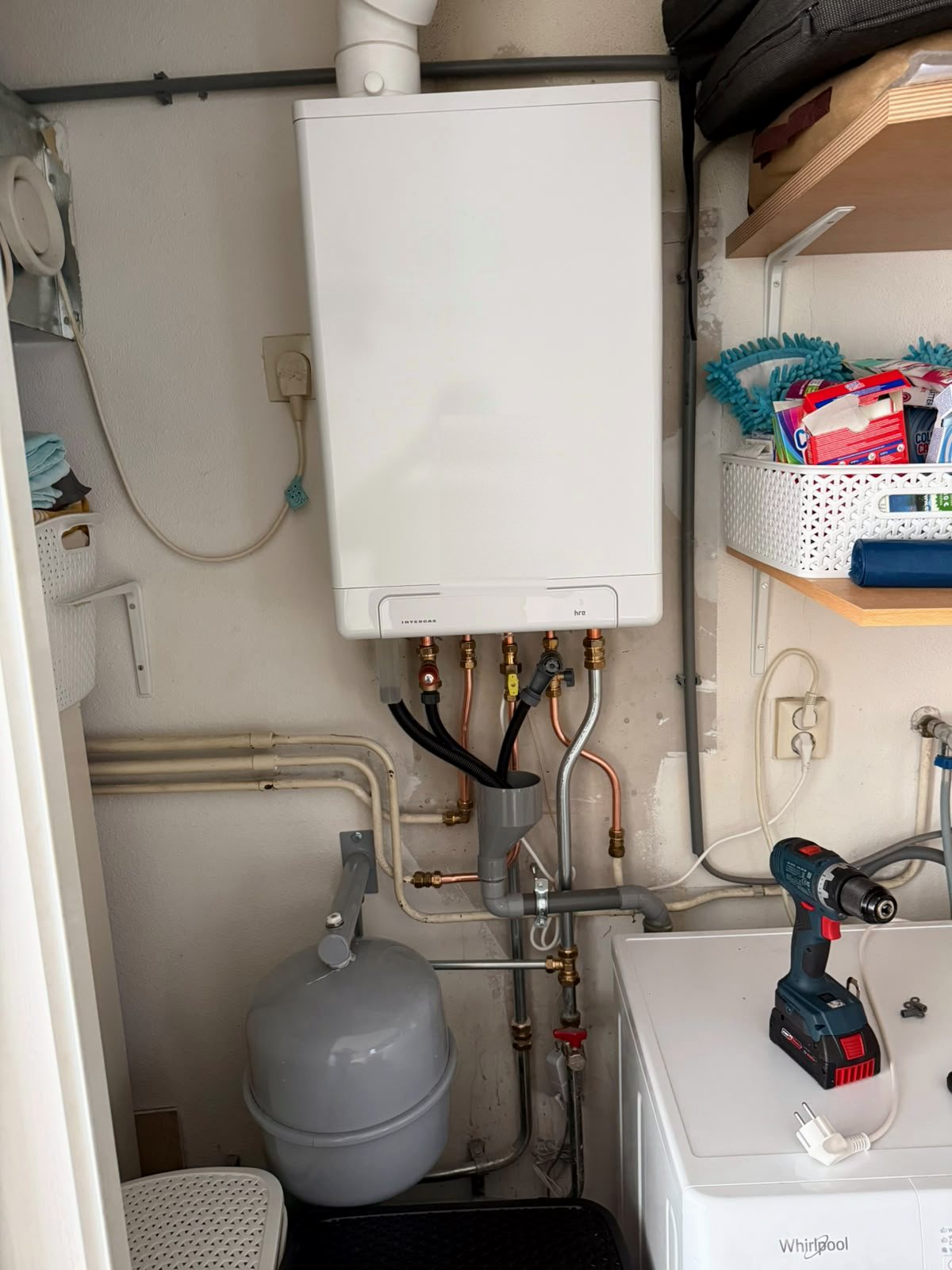 Intergas cv-ketel aan de muur in een bijkeuken boven een wasmachine, met koperen aansluitingen en een expansievat op de vloer