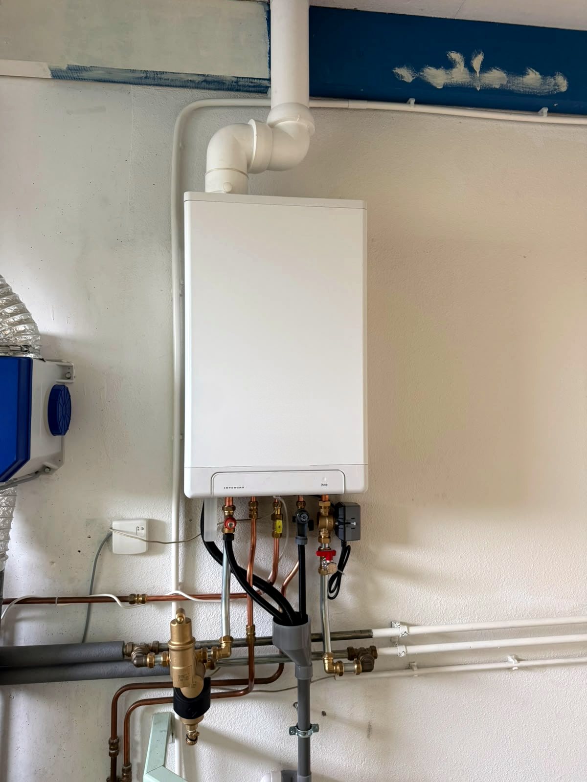 Intergas cv-ketel tegen een witte wand met koperen en stalen leidingen die horizontaal verder lopen langs de muur