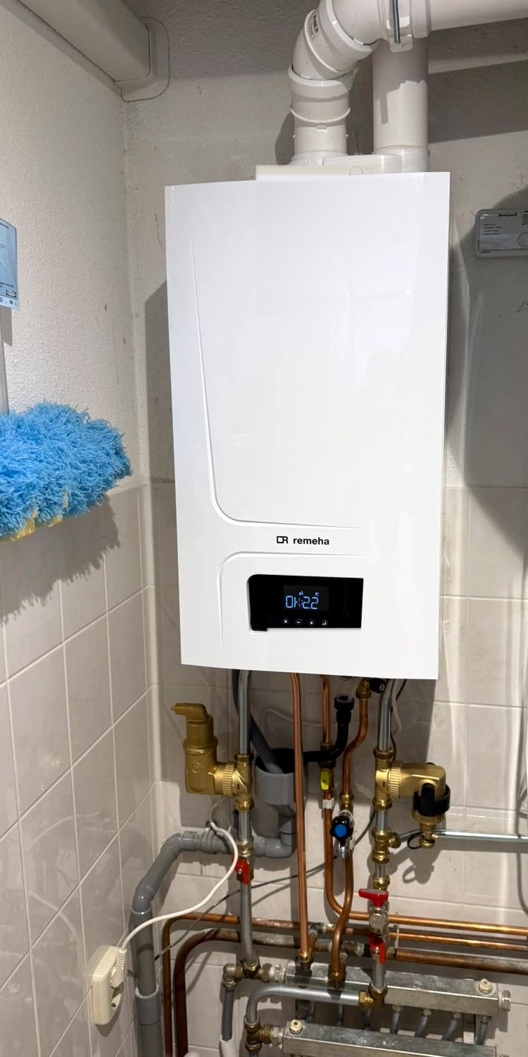 Remeha cv-ketel tegen een betegelde wand met daaronder koperen leidingen en een verdeler voor vloerverwarming